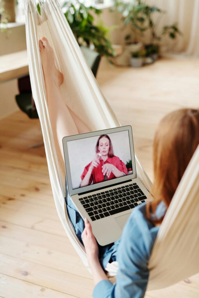 A woman relaxes in a hammock while having a video call on her laptop indoors.